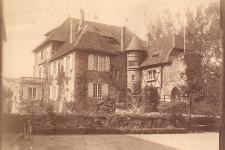 The photo shows Lede Castle around 1900 in an old sepia photograph. There is architecture with a turret placed between two sides of the building, a bay window with a balcony above, a castle gate and shutters.